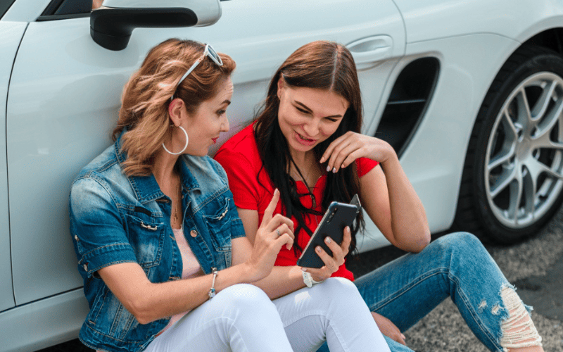 Cual es el precio de incluir a tu hijo en el seguro de coche. Imagen de unas jóvenes consultando su móvil apoyadas en el coche.